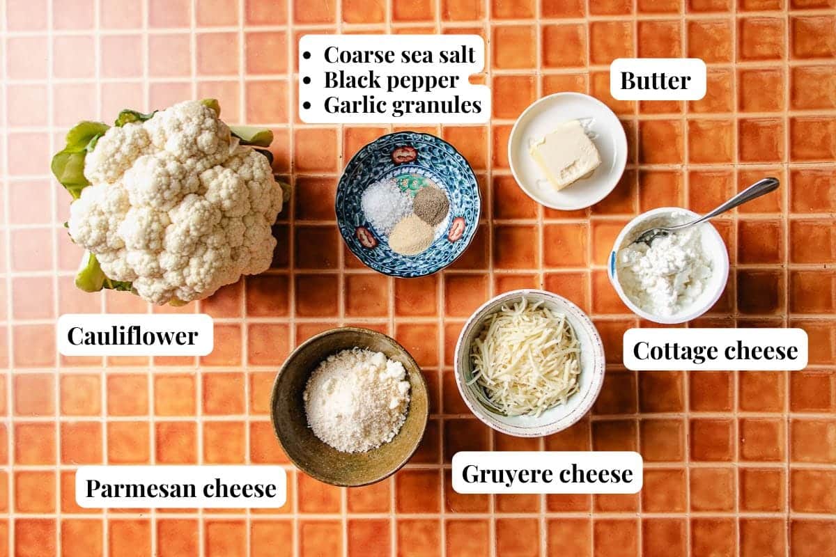 A flat lay of labeled ingredients for a Low Carb Cauliflower Mash on an orange tile surface: cauliflower, Parmesan cheese, Gruyere, cottage cheese, butter, and a bowl with coarse sea salt, black pepper, and garlic granules.