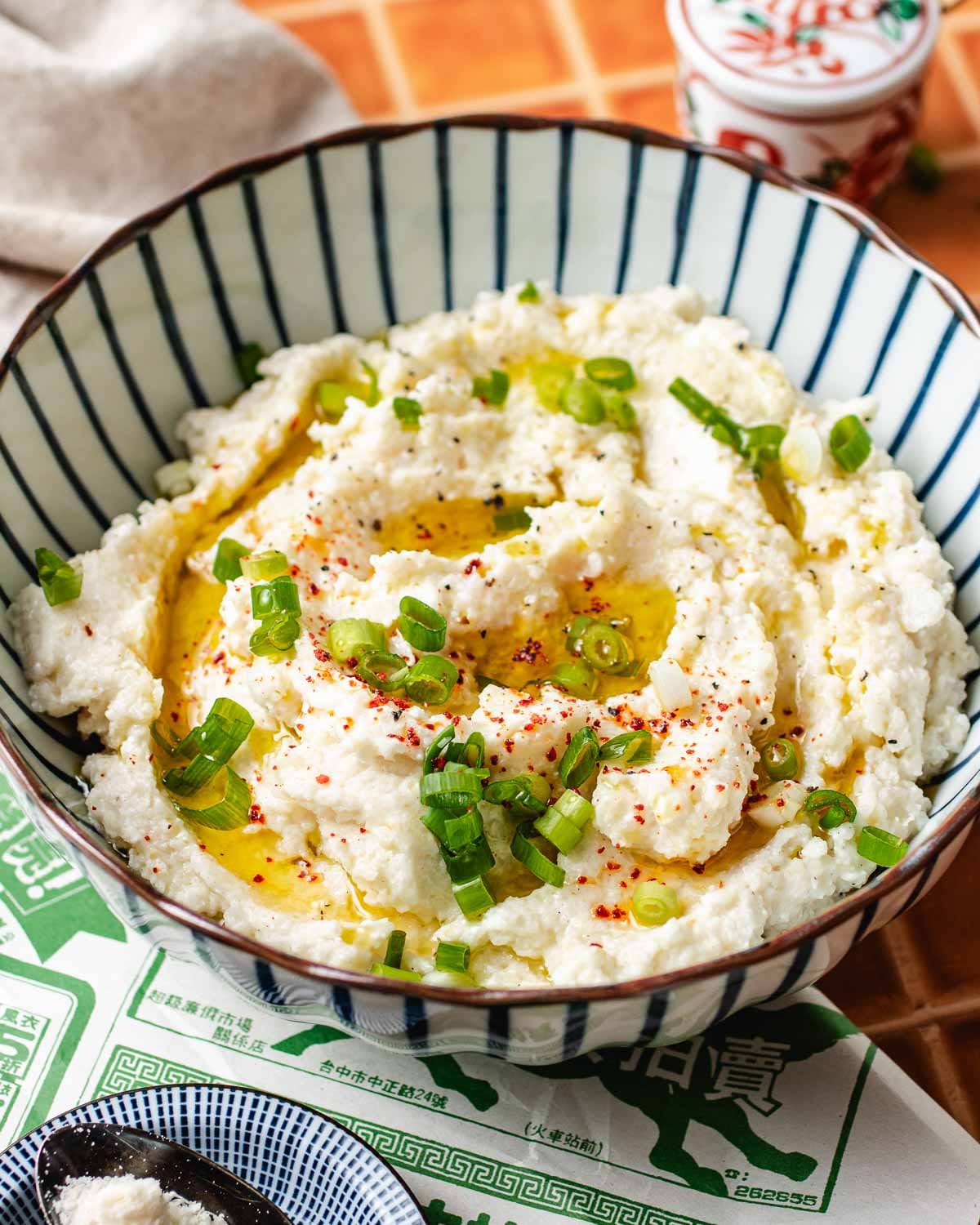 A bowl of creamy cauliflower mashed potatoes topped with olive oil, chopped green onions, and a sprinkle of red chili flakes, served in a blue-striped bowl on a patterned placemat.