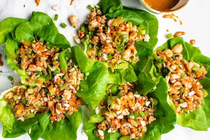 Five healthy lettuce wraps filled with chopped vegetables, seasoned ground chicken, and topped with nuts and green onions, arranged on a white surface with a bowl of sauce nearby.