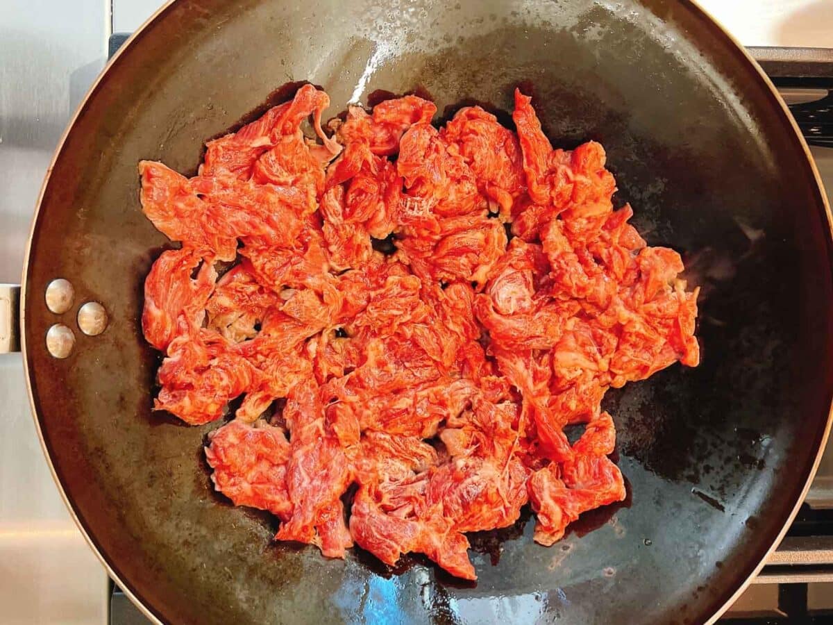 Thin slices of raw velveted beef, prepared using the velvet beef for tender stir fry method, are spread out in a large, black frying pan on a stovetop, ready to be cooked.