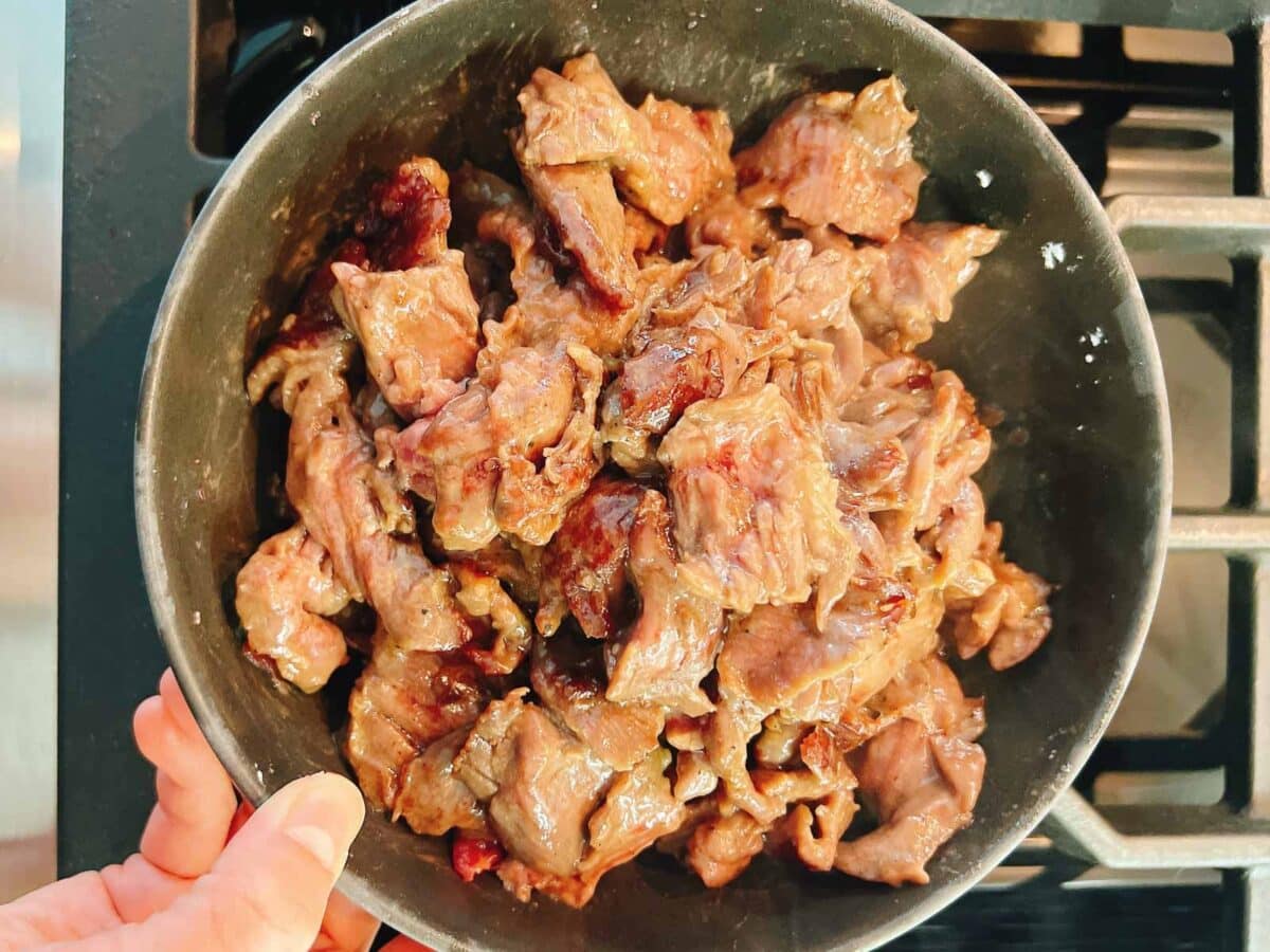 A hand holds a black bowl filled with pieces of tender and juicy cooked, browned steak next to a stove burner—showcasing how to velvet beef for tender stir fry.