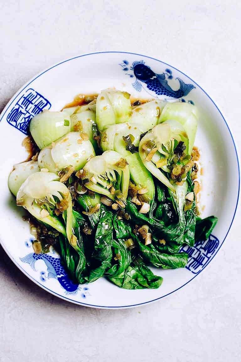 Photo shows halved baby bok choy blanched and drizzled with Chinese ginger garlic dim sum sauce over a white blue plate. 