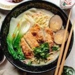 A bowl of creamy chicken ramen topped with sliced fried chicken, two halves of a marinated boiled egg, bok choy, and chopped green onions, served with chopsticks.