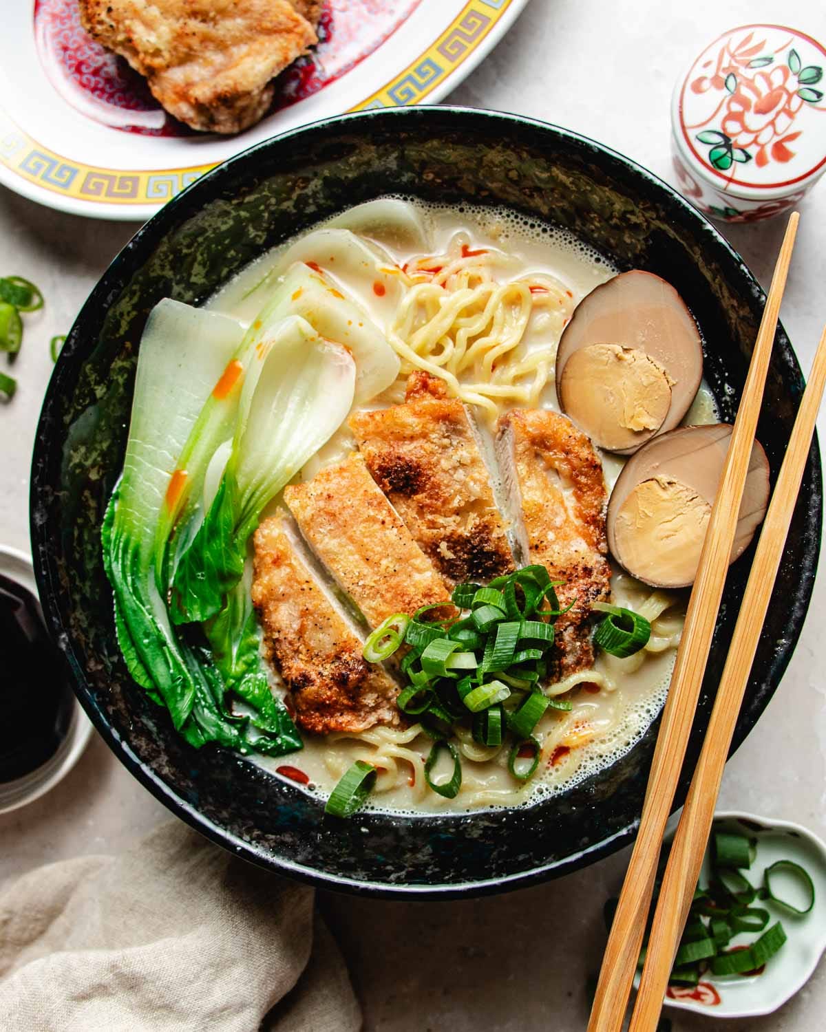 A bowl of chicken ramen noodle soup in a creamy broth topped with sliced crispy chicken, two halves of a soy-marinated egg, bok choy, and chopped scallions, with chopsticks resting on the side.