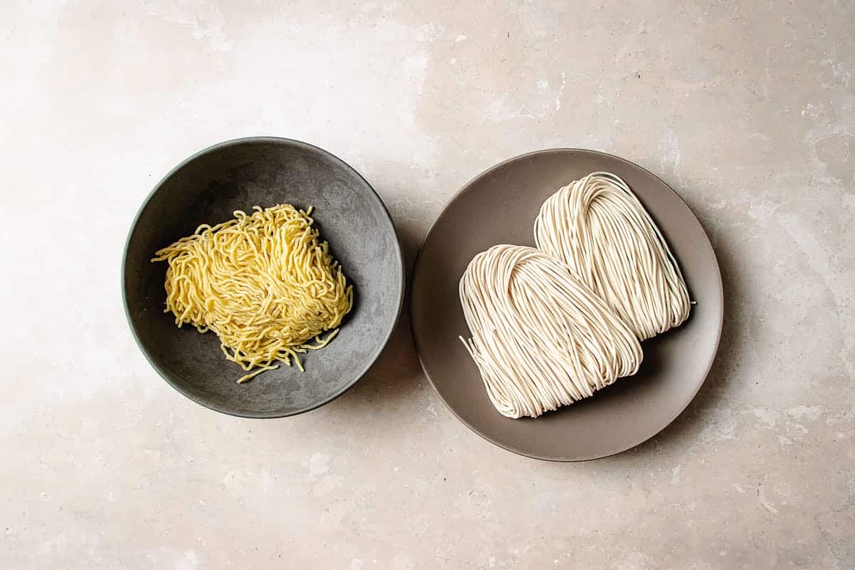 A dark plate of fresh ramen with thin wavy yellow noodles next to a plate with two bundles of folded dry wheat ramen noodles.