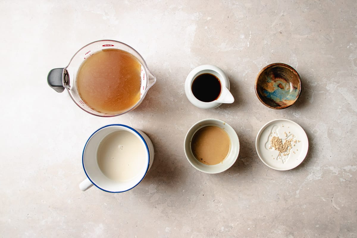 A glass measuring cup of chicken broth next to bowls containing soy sauce, tahini, a small amount of sesame seeds, and a cup of plain soy milk for building ramen broth.