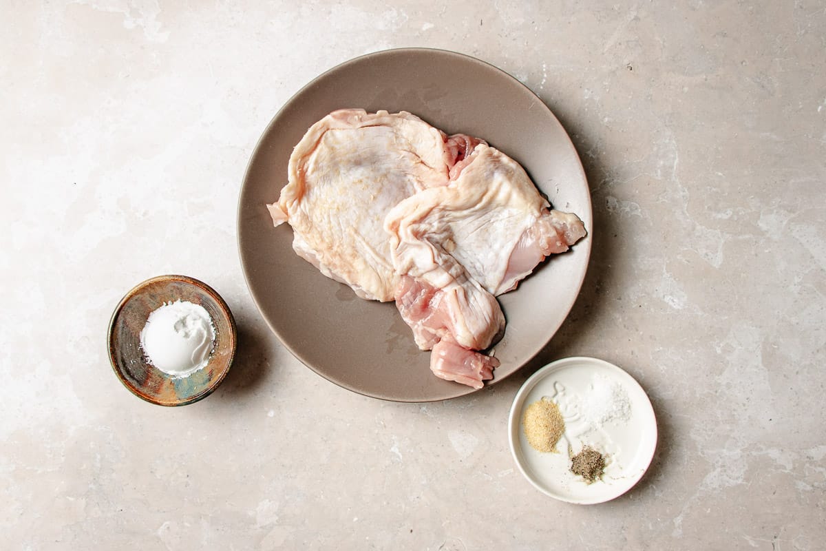 Two raw boneless and skin on chicken thighs for chicken ramen on a plate, next to a small bowl of white powder and a small dish of spices.
