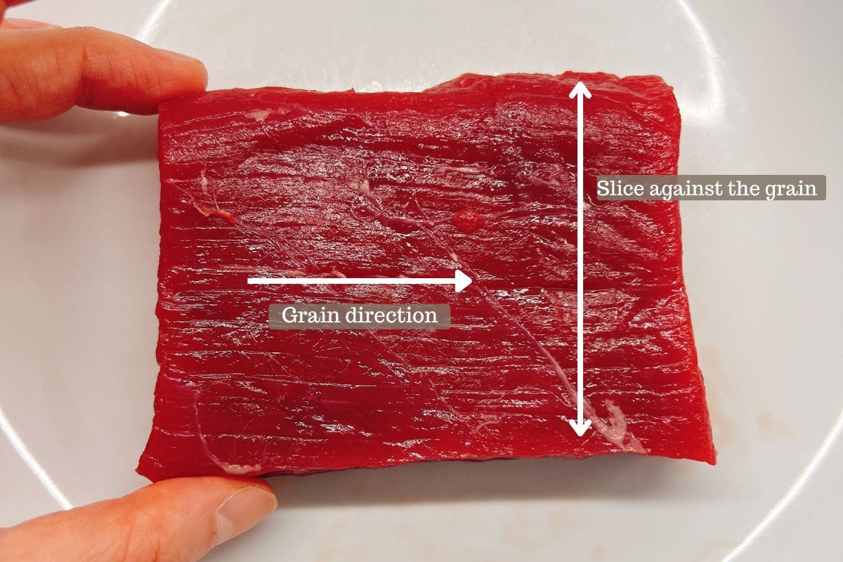 A piece of raw red meat on a white plate, held by two fingers, with arrows showing the grain direction (horizontal) and where to slice against the grain (vertical)—an essential tip when choosing the best beef cut for stir fry.