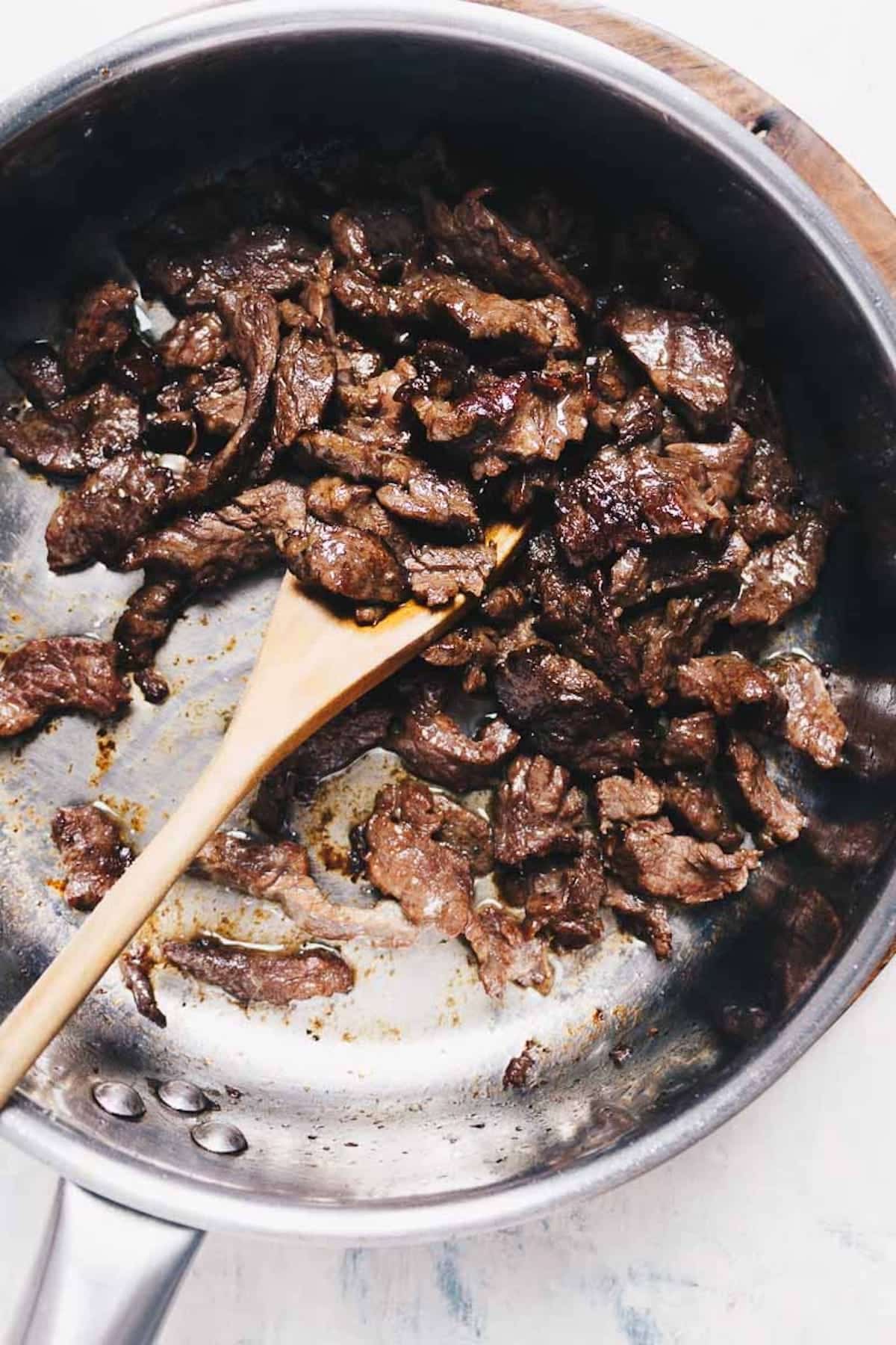 Slices of stir fried beef in a stainless steel pan, being stirred with a wooden spoon. The beef is browned, well-seasoned, and looks like the best velveted beef for stir fry—perfectly velvet beef for tender stir fry dishes.