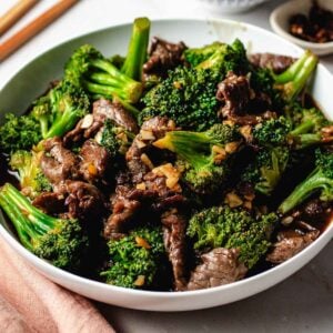 A white bowl filled with beef and broccoli stir-fry, featuring velvet beef for tender stir fry, bright green broccoli florets, and chopped garlic in a savory sauce. Chopsticks and a napkin are placed nearby.
