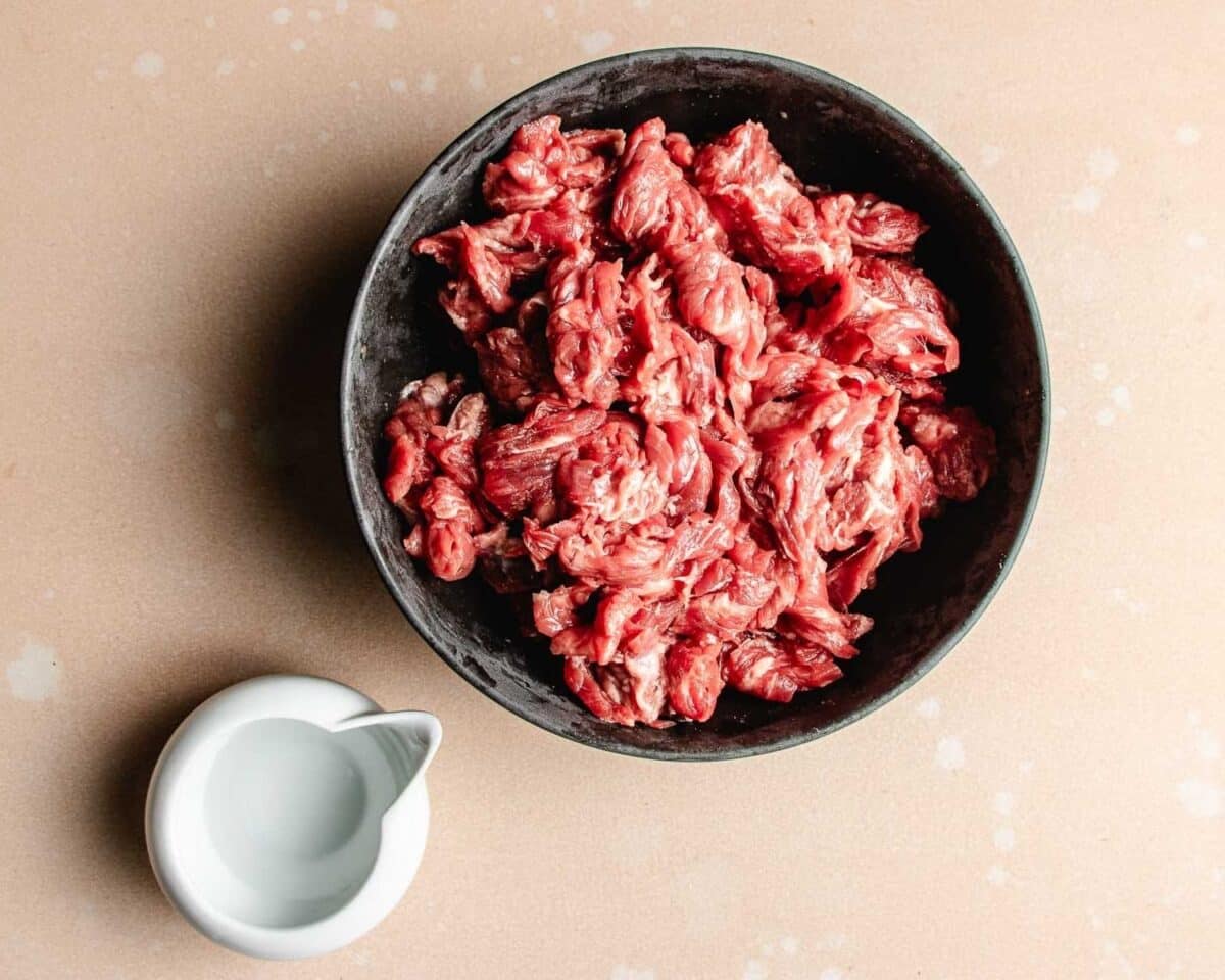 A black bowl filled with raw, sliced beef—perfect for Velvet beef for tender stir fry—sits on a light beige surface next to a small white pitcher of water to rehydrate the beef for tender velveting texture.