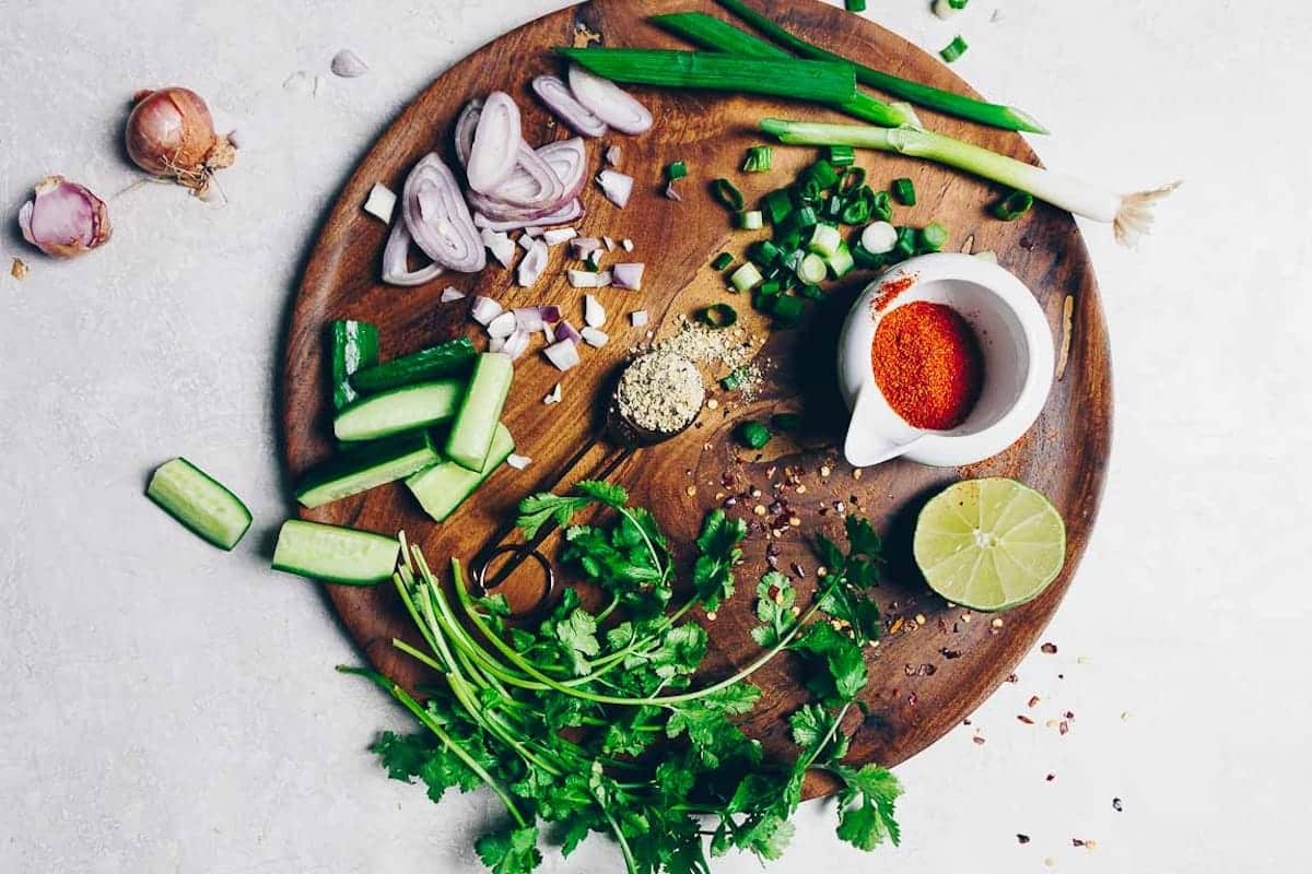 Round wooden board with chopped cucumber, cilantro, green onions, sliced shallots, minced garlic, lime half, spices in a small white bowl, and red pepper flakes on a light surface—perfect for preparing authentic Thai larb or classic larb moo.