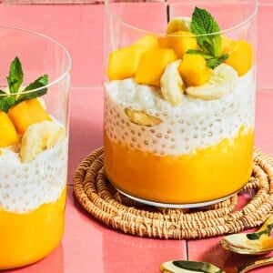 Image shows 2 glasswares filled with mango puree, sago pudding, and chopped mango and mint leaves served as a dessert.