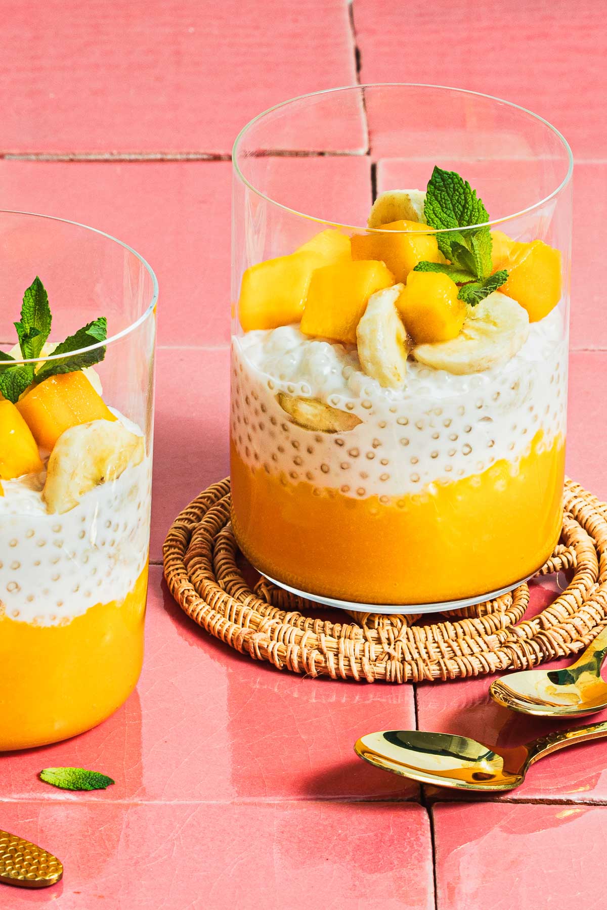 Image shows 2 glasswares filled with mango puree, sago pudding, and chopped mango and mint leaves served as a dessert.