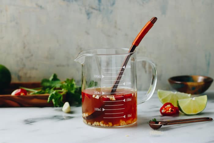 A glass measuring cup filled with a red Vietnamese dipping sauce (nuoc cham) and chopped garlic sits with a spoon inside. Fresh herbs, chili, garlic, and lime wedges rest on the marble surface nearby, with a wooden bowl in the background.