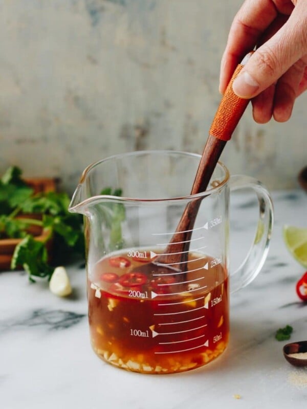 A hand stirs Vietnamese dipping sauce (nuoc cham), with chopped garlic and red chili slices in a glass measuring cup on a marble surface, surrounded by herbs, lime, and garlic.