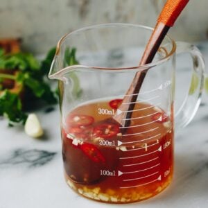 A glass measuring cup filled with Nuoc Cham—a classic Vietnamese Dipping Sauce featuring brownish liquid, chopped garlic, and red chili, with a wooden spoon inside. Fresh cilantro and garlic cloves are blurred in the background.