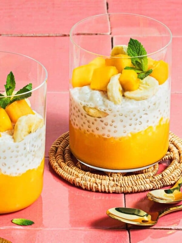 Image shows 2 glasswares filled with mango puree, sago pudding, and chopped mango and mint leaves served as a dessert.