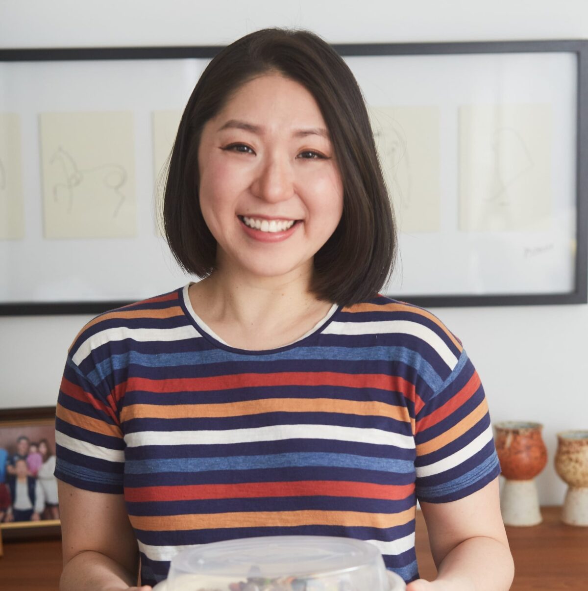 ChihYu Smith with shoulder-length dark hair, wearing a striped shirt, smiles at the camera while holding a container of Creamy Roasted Sesame Dressing. Framed drawings and a family photo are visible on the wall and table behind her.