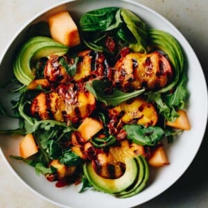 Recipe image shows grilled peaches served on a big white plate with salad greens, avocado, and prosciutto and balsamic sauce on top.