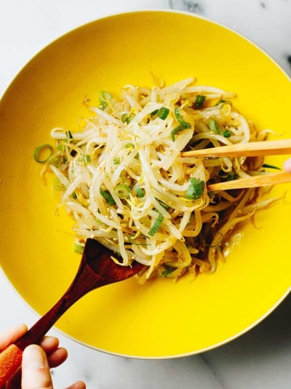 A close overhead shot shows blanched bean sprouts mixed with sauce Korean style, served in a bright yellow bowl