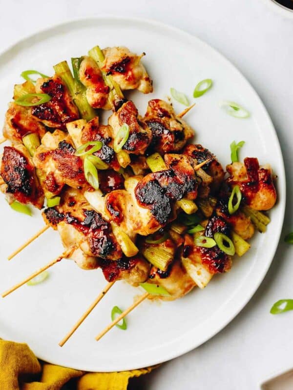 Negima yakitori skewers garnished with sliced green onions, served on a white plate. Some skewers have char marks, and a few green onion slices are scattered around the plate.
