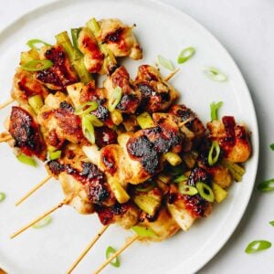 Japanese chicken Yakitori skewers with charred edges and green onions on a white plate, garnished with sliced scallions.