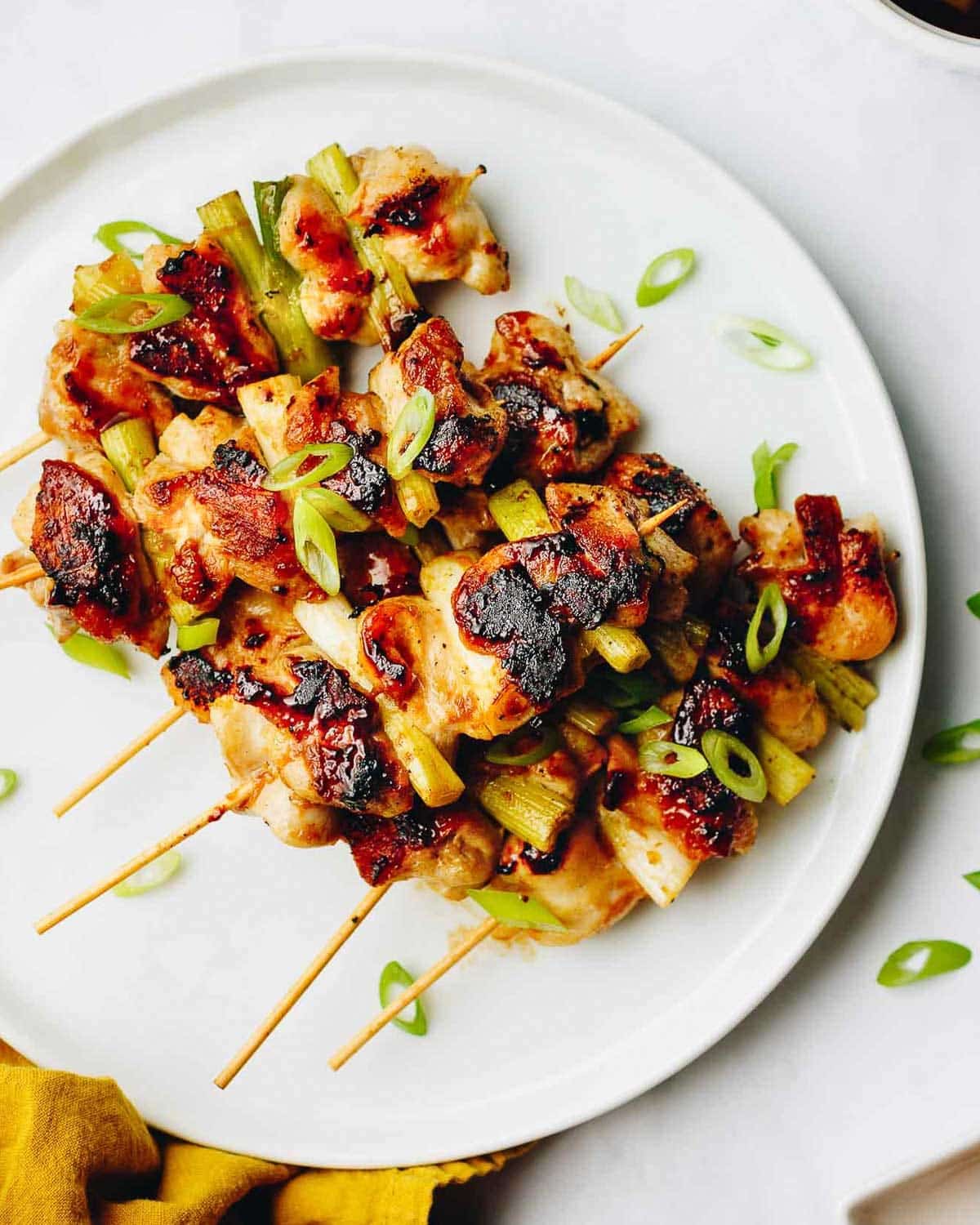 Negima yakitori skewers garnished with sliced green onions, served on a white plate. Some skewers have char marks, and a few green onion slices are scattered around the plate.