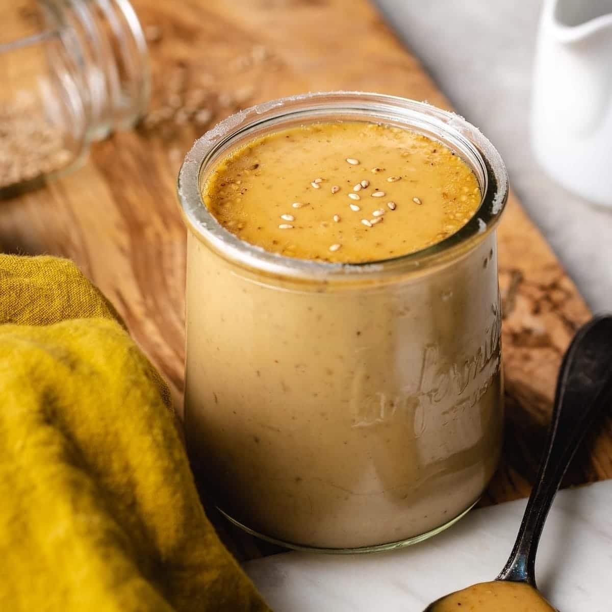 A glass jar filled with creamy brown tahini, reminiscent of Creamy Roasted Sesame Dressing, sits on a wooden surface beside a yellow cloth and a spoon with tahini. An open jar and a white pitcher are in the background.