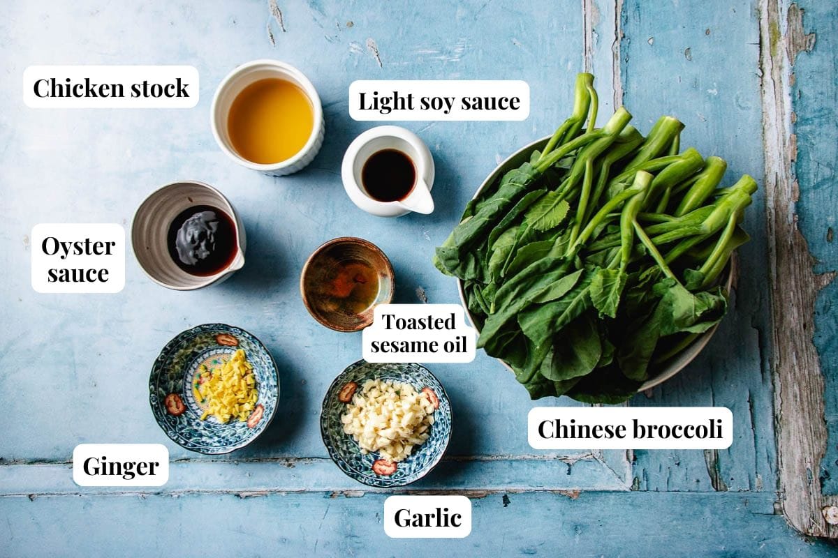 A flat lay of labeled ingredients for Chinese Broccoli with Oyster Sauce, including chicken stock, light soy sauce, toasted sesame oil, ginger, garlic, and a bowl of fresh Chinese broccoli on a blue wooden surface.