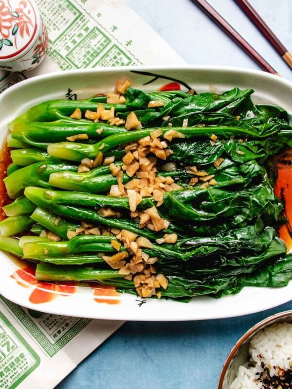 A rectangular plate of Chinese broccoli (gai lan) topped with chopped garlic and a savory drizzle of Broccoli Chinese Oyster Sauce, set on a light surface with chopsticks and a bowl of rice nearby.