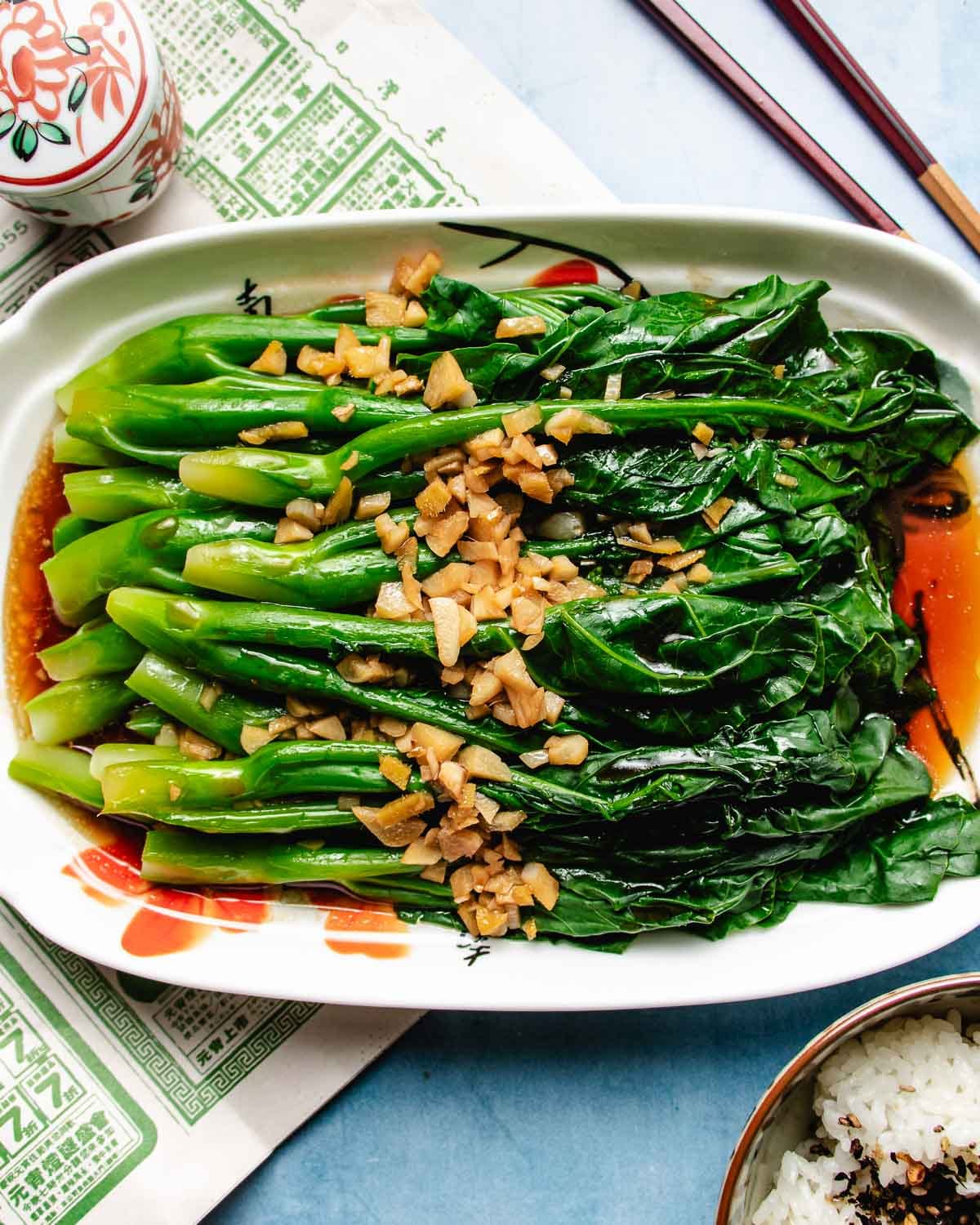 A rectangular plate of Chinese broccoli (gai lan) topped with chopped garlic and a savory drizzle of Broccoli Chinese Oyster Sauce, set on a light surface with chopsticks and a bowl of rice nearby.