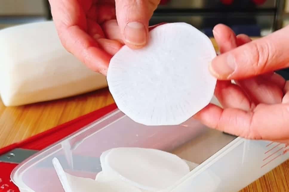 A person holds a thin, translucent slice of white radish above a container with more slices on a wooden surface, suggesting a creative wrapper alternative for low carb dumplings like Chinese jiaozi. A whole radish and slicer are nearby.