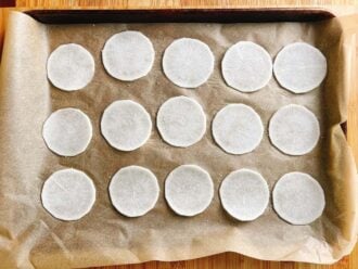 A baking tray lined with parchment paper holds fifteen evenly spaced, thin, round slices of white daikon radish—perfect as low carb dumplings wrappers—arranged in three neat rows on a wooden surface.