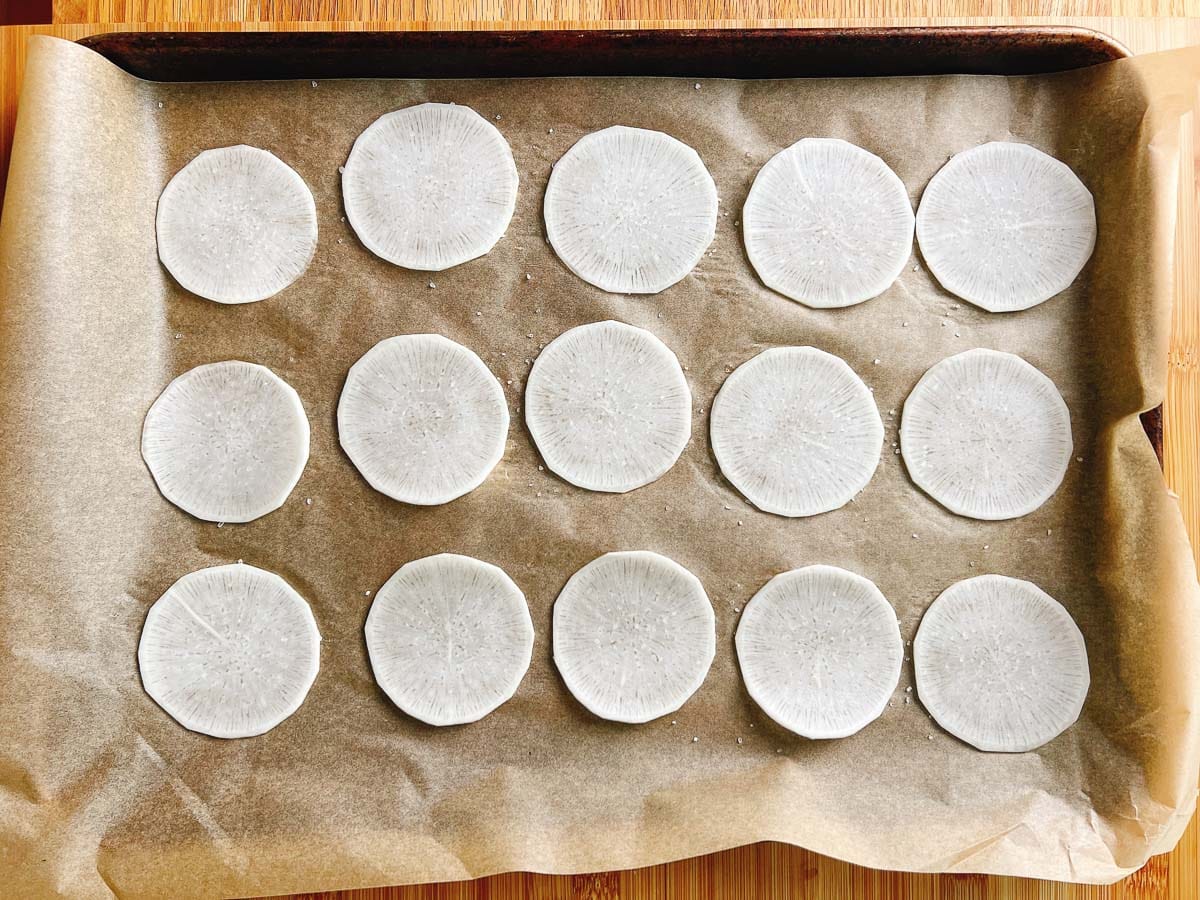 A baking tray lined with parchment paper holds fifteen evenly spaced, thin, round slices of white daikon radish—perfect as low carb dumplings wrappers—arranged in three neat rows on a wooden surface.