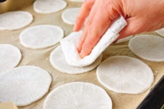 A hand uses a paper towel to pat dry thinly sliced white radishes arranged on parchment paper, preparing them as low carb dumplings wraps for homemade jiaozi.