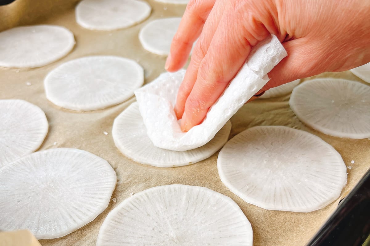A hand uses a paper towel to pat dry thinly sliced white radishes arranged on parchment paper, preparing them as low carb dumplings wraps for homemade jiaozi.
