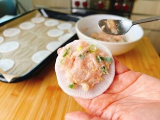 A hand holds a thin white radish slice topped with raw ground meat and chopped green onions—an inventive twist on low carb dumplings. In the background, a bowl of filling and a tray lined with more radish slices await assembly.