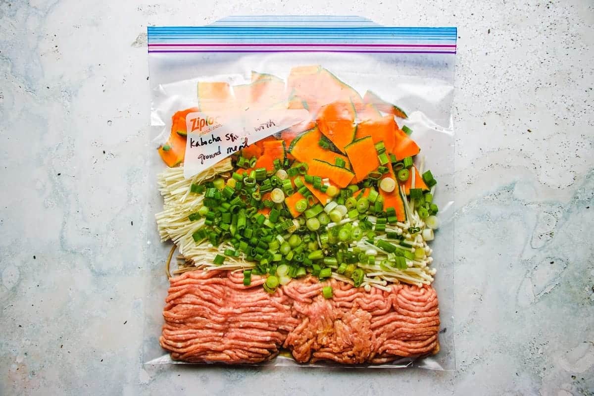 A storage bag with labeled contents: sliced orange kabocha pumpkin, chopped green onions, enoki mushrooms, and ground chicken, arranged in layers on a light surface—perfect for your next Kabocha Recipe.