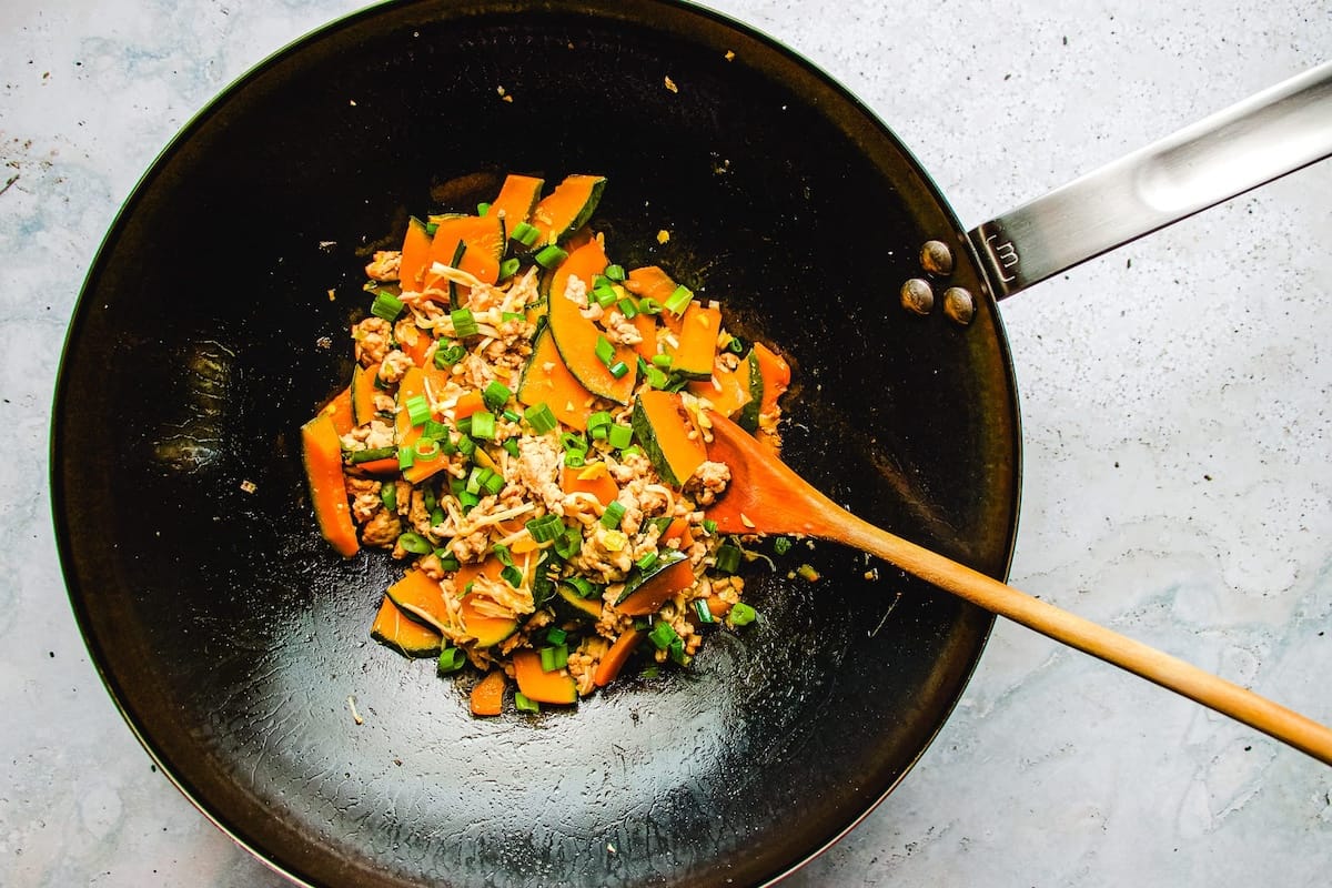 A black wok with ground chicken, Japanese pumpkin, and herbs being simmered with a wooden spoon, on a light-colored countertop.