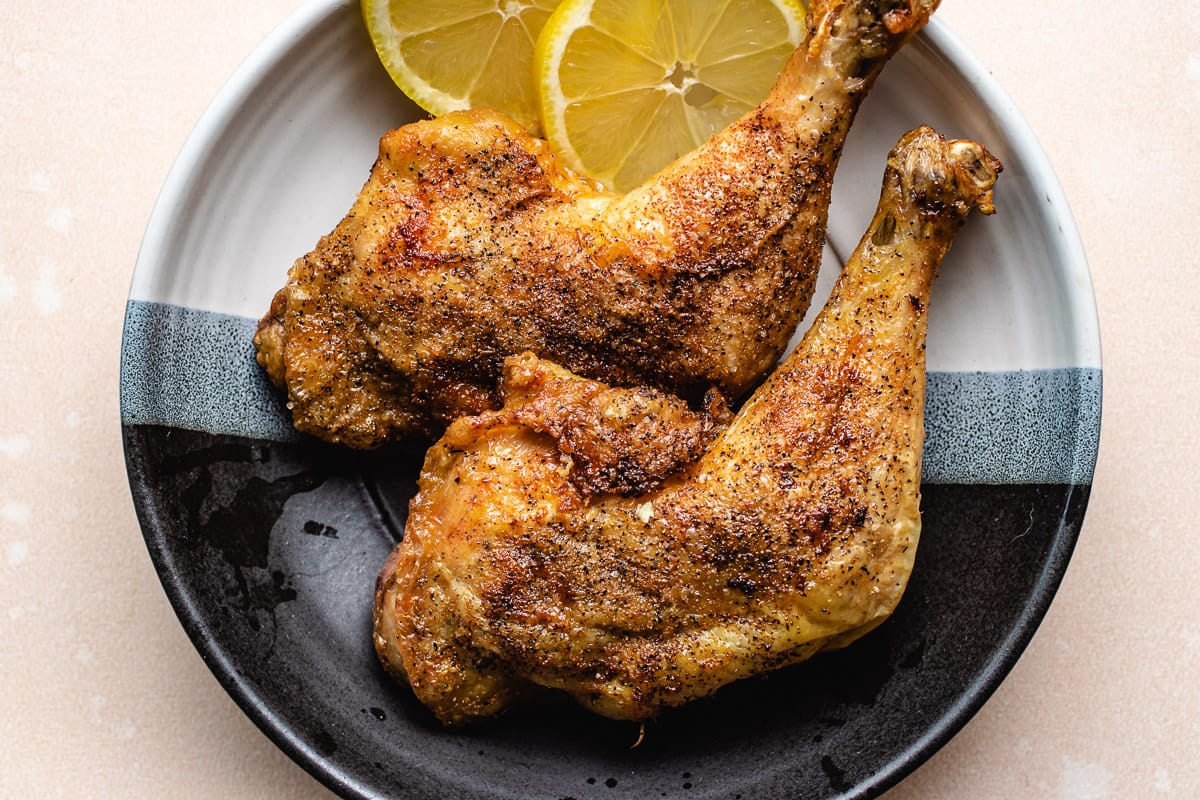 Two seasoned and roasted chicken leg quarters on a black and white plate, garnished with two lemon slices—perfect for high protein low carb recipes or low carb meal prep ideas.