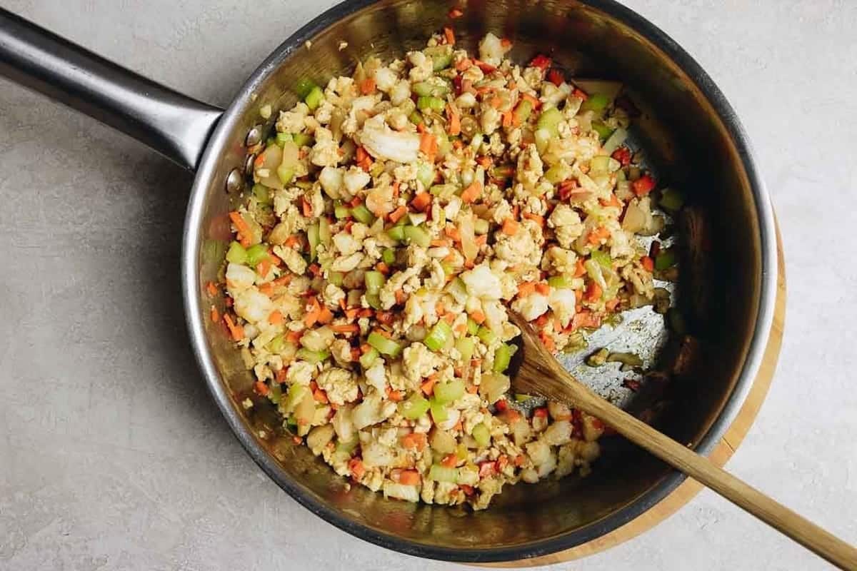 A stainless steel pan with chopped vegetables and ground chicken being cooked, including carrots, celery, and ground chicken, is stirred for healthy lettuce wraps on a light-colored countertop.