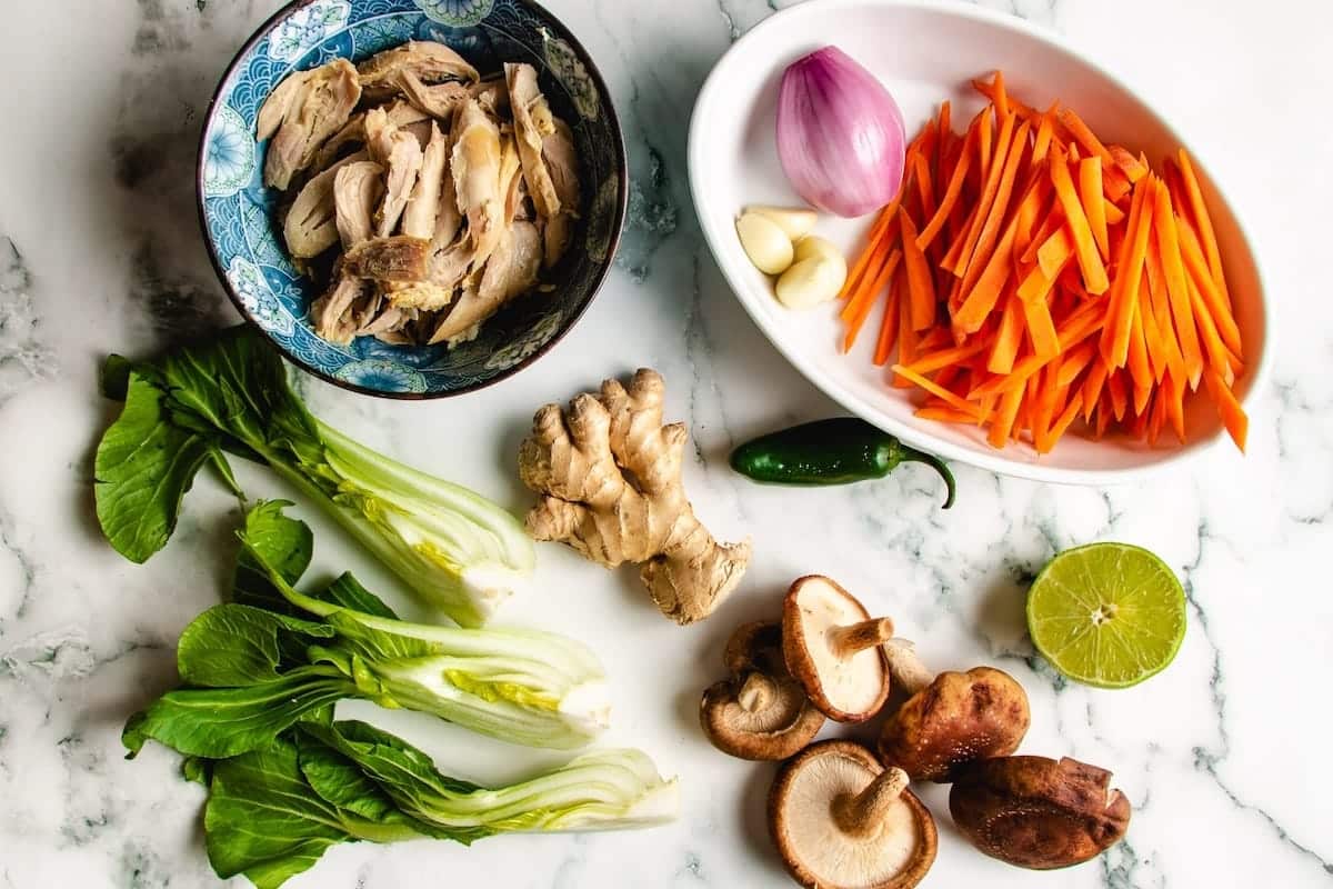 Ingredients on a marble surface, ready for Asian Chicken Noodle Soup: bok choy, shredded chicken, sliced carrots, shallot, garlic cloves, ginger root, shiitake mushrooms, a green chili, and half a lime.