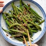 A plate of Chinese garlic green beans topped with sesame seeds and small bits of garlic, served in a blue and white patterned bowl, with chopsticks picking up some beans.