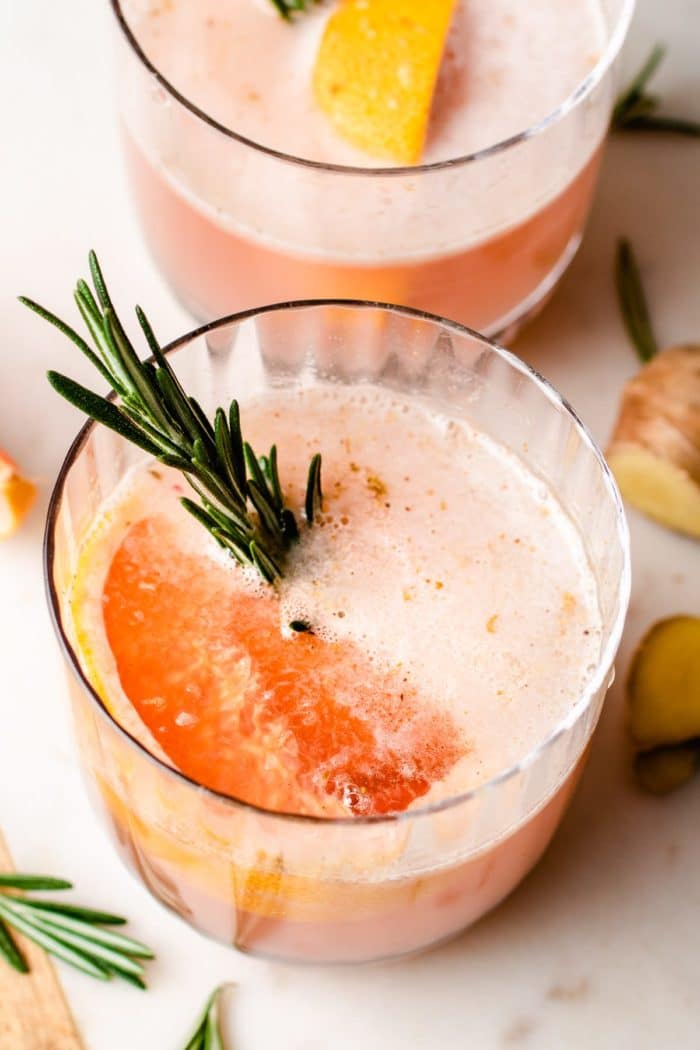 Photo shows 2 cocktail glasses with frothy sake and a piece of grapefruit with rosemary in the glass.