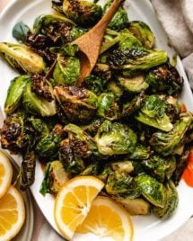 Photo shows a white plate loaded with crispy air fried brussels sprouts glazed with maple lemon sauce and with lemon on the side