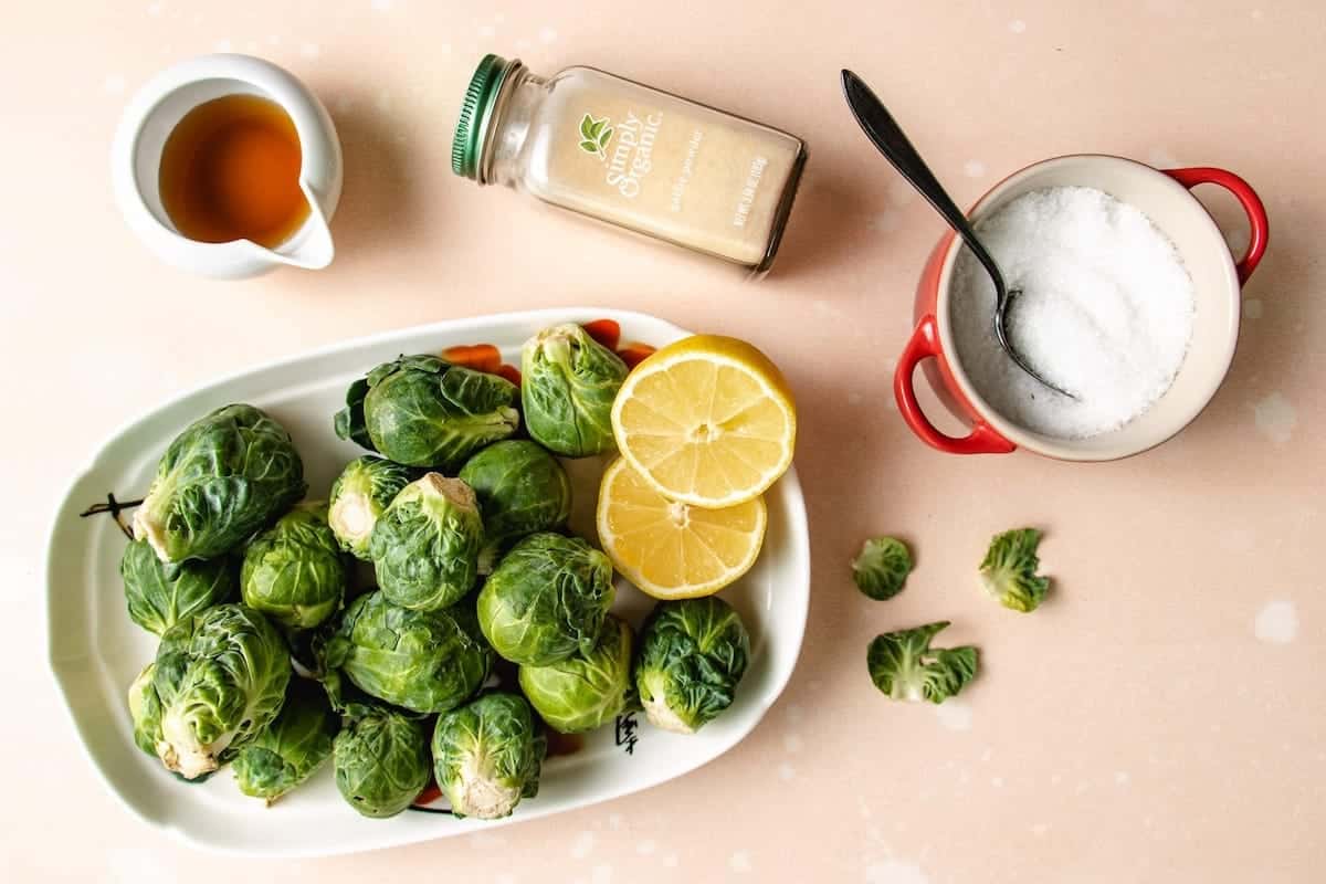 Brussels sprouts on a white platter, halved lemons, a small pitcher of maple syrup, a jar of garlic granules, and a red bowl of salt with a spoon sit atop a light pink surface—perfect for preparing delicious maple glazed brussel sprouts.