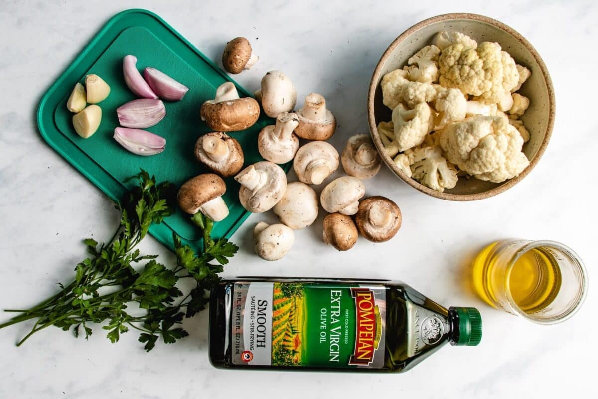 A cutting board with garlic, shallots, parsley, mushrooms, a bowl of cauliflower florets, a jar of olive oil on a white surface—perfect for preparing your favorite sauteed cauliflower and mushrooms or trying out a new mushroom cauliflower recipe.