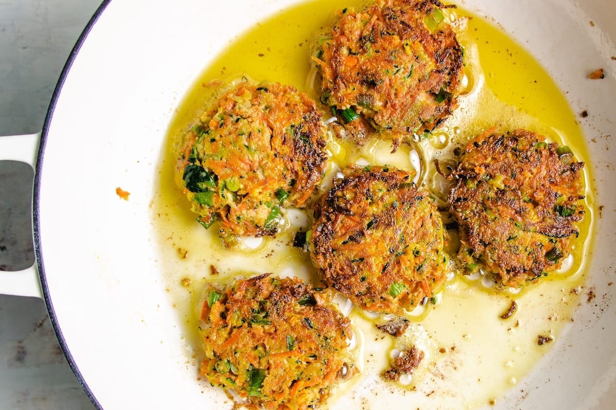 Six golden-brown Zucchini Carrot Fritters are frying in oil in a white skillet, with visible grated carrots, green herbs, and some browning on the edges.