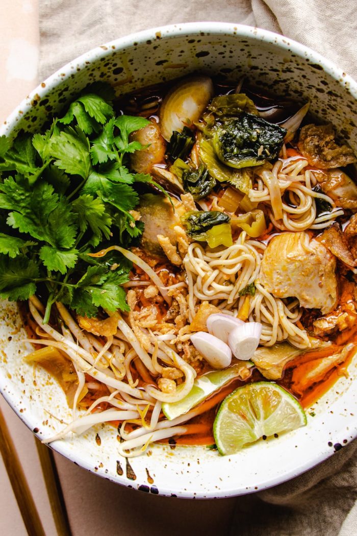 Feature image shows a big white noodle soup bowl with curry chicken, noodles, lime, and cilantro in a creamy chiang mai noodle curry khao soi gai style broth.